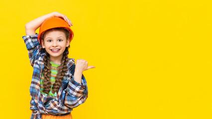 Fototapeta A little girl in a hard hat points to your advertisement on a yellow isolated background. A child in the clothes of a worker, preparing for repairs. Copy space. Banner. A place for advertising. obraz