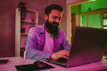 A happy IT specialist is typing on his laptop at night. Neon purple lighting is on.