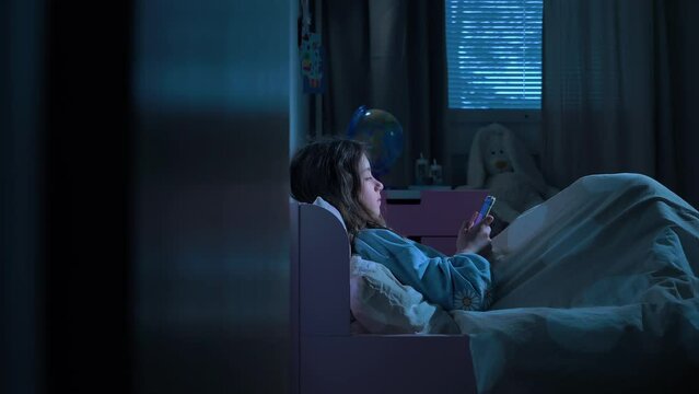 Teen girl scrolling social media texting on smartphone in bed in bedroom at night. Technology, internet, communication and people concept. Dependence on social networks in children. Not healthy sleep.