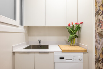 Close-up of a sink on a kitchen surface with cabinets and a vase of tulips in a modern stylish white kitchen. The concept of a stylish and comfortable kitchen in a small apartment. Copyspace