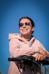 Fototapeta na wymiar A woman in a pink sweatshirt and blue jeans stands next to a bicycle. Sports and healthy lifestyle.