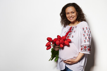 Authentic portrait of a curly haired charming brunette, pregnant woman touching her belly, smiling looking at camera, holding bunch of red tulips, dressed in ukrainian ethnic dress, white background