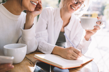 Female colleagues creating content information during collaboration meeting for discussing business ideas together, cheerful women with coffee cup talking about brainstorming in coworking