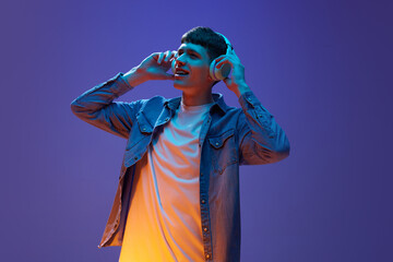 Fototapeta premium Portrait of young relaxed man in casual clothes, listening to music in headphones, having fun against gradient purple background in neon light. Concept of human emotions, lifestyle, youth