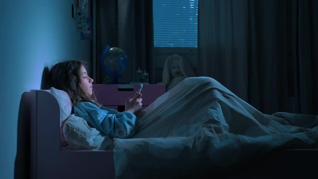 Teen girl lying in bed at night using smartphone, scrolling social media and texting. Not healthy sleep. Insomnia and the harm of the smartphone before going to bed. Dependence on social networks