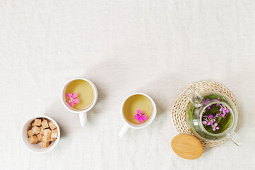 Fireweed tea in white cups and transparent glass teapot, herbal hot tea from green leaves of ivan chai on textile tablecloth. Top view healthy drink and wild flowering willow-herb, tea time