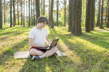 Young man is sitting on the grass in park with his laptop. Distance learning concept.