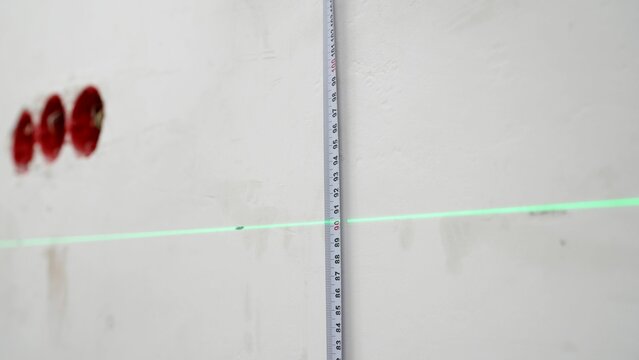 A man uses a cross laser level and a tape for accurate measurements on a light wall, close-up. Measure the height with a laser level. Laser level and tape measure in action.