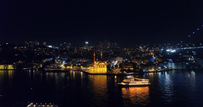 Aerial view of Ortakoy Mosque and flying over an illuminated tour boat at night. Buyuk Mecidiye Camii well known as Ortakoy Mosque and it's located in an amazing place by the waters close to Bosphorus