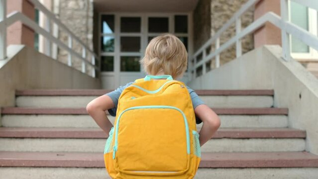 Little schoolboy with a backpack goes to school after summer holiday. Student going up the stairs of school building. Kids back to school concept.