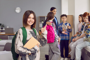 Fototapeta premium Portrait of happy girl school student showing thumb up. Beautiful smiling schoolgirl wearing casual clothes standing with backpack on background of her classmates in classroom and looking at camera