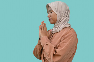 Portrait of a young beautiful southeast Asian Muslim girl wearing a hijab gesturing Eid Mubarak greeting on blue background. closing her eyes while praying.  