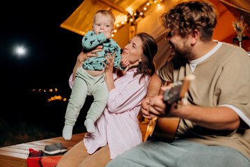 Happy family relaxing and spend time together in glamping on summer evening and playing guitar near cozy bonfire. Luxury camping tent for outdoor recreation and recreation. Lifestyle concept - 601148395