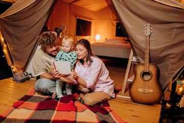 Happy family with lovely baby playing and read book together in glamping on summer evening. Luxury camping tent for outdoor recreation and recreation. Lifestyle concept