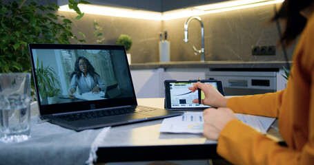 Unknown modern dark-haired woman in stylish clothes sitting in front of computer during video call with african american female colleague at home and talking about business strategy