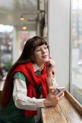 Attractive young woman drinking coffee in modern cafe, using mobile phone, looking away