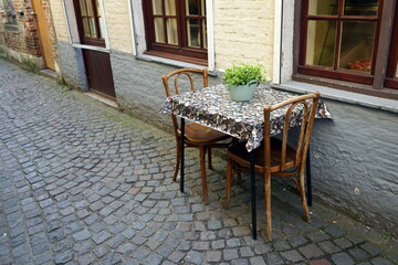 Einsamer Tisch für Zwei vor einem Restaurant und Bistro als Teil des Unesco Weltkulturerbe im Frühling mit Grünpflanze als Tischdeko in den Gassen der Altstadt von Brügge in Westflandern in Belgien