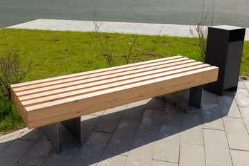 Illuminated by sunlight, a new wooden bench and a metal trash can outdoor on the background of a...
