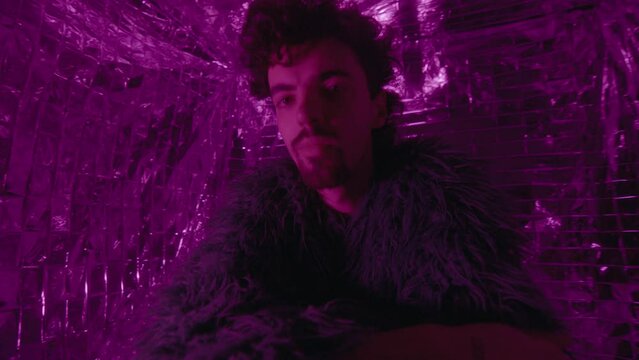 Male fashion model wearing stylish feather jacket and pearl necklace posing on camera in colorful neon light against silver background. Medium shot, creative portrait