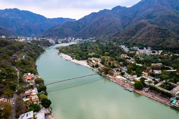 Wall murals Himalayas aerial drone shot of blue water of river ganga stretching into distance with himalayas with ram setu suspension bridge and temples on the banks of the river