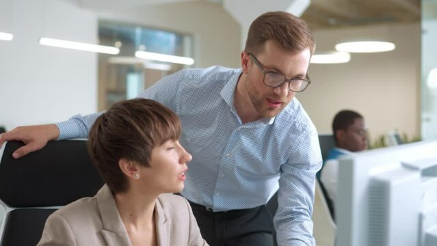 Caucasian workers communicating in front of computer. Attractive leader controlling and looking at work results of his beautiful assistant. Pretty woman listening with focus to his advices.