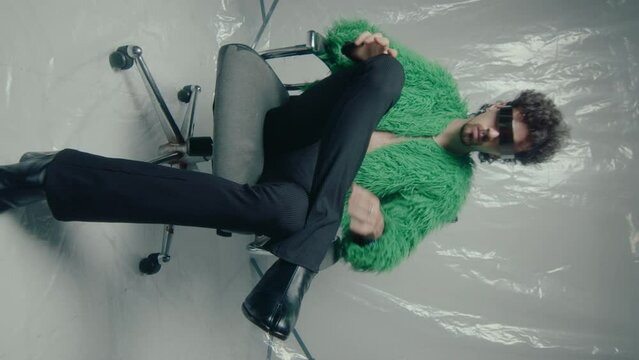 Attractive man wearing feather jacket, tabi boots, bell-bottoms, trendy sunglasses sitting on chair in studio, posing on plastic wrap backdrop during photoshoot. Vertical format video, zoom shot