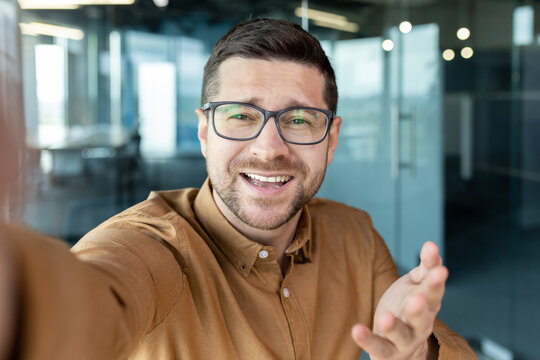 Close-up photo. Young man freelancer, programmer takes a selfie on the phone and talks on a video call in the office. He holds a device in his hand, smiles at the camera