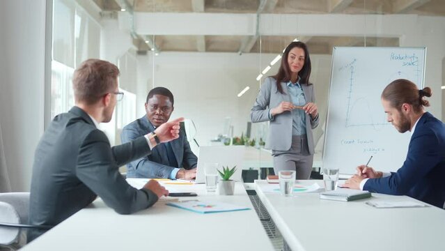 Mixed-raced managers meeting, brainstorming ideas for business project. Young woman standing near white board while coworkers sitting at table and communicating in conference room.