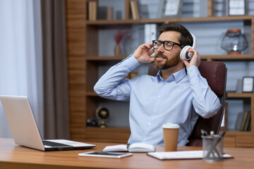 Rest, break at work. A young businessman sitting in the office, leaning on a chair, listening to music in headphones, closed his eyes, relaxed.