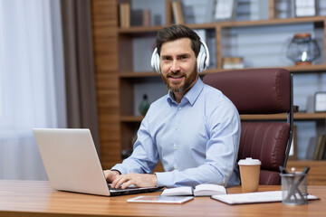 Portrait of a young male businessman working in the office at a laptop, sitting in white headphones and smiling at the camera.