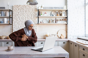 Heart attack, stroke at home. A young Muslim woman in a hijab works at home in the kitchen on a...