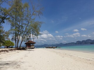 Beach rocks. Two boats on the shore near the pavilion for rest. Trees provide shady and shade. Green sea, white sand, blue sky with light clouds. Islands and mountains in background.