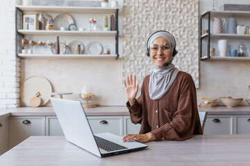 Young Muslim woman in hijab and headset, blogger doing remote online broadcast, communicating via video call. Sitting at home in the kitchen at the laptop. A smiling woman waves hand at the camera.