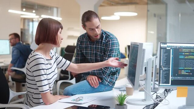 Handsome Caucasian programmer sitting on table and supporting or helping beautiful female developer. Busy people communicating about creating websites for business company. Partnership concept.