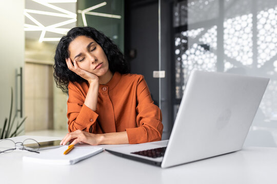 Beautiful tired hispanic woman sleeping on desk, close-up business woman with closed eyes napping inside office near laptop.