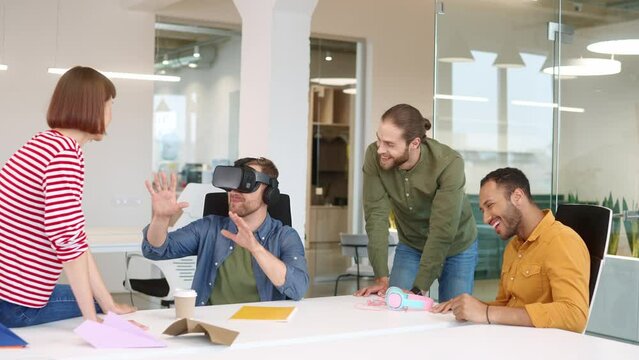 Group of mixed-raced people sitting in office and communicating with each other. Funny man wearing virtual reality headset and checking look of future office. Workers smiling and laughing together.
