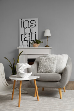 Cozy grey armchair with cushions, decorative figure on table and fireplace in interior of living room