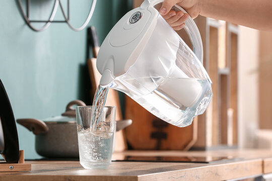 Woman pouring water from modern filter jug into glass on kitchen counter