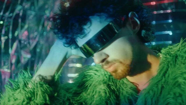 Glamorous man wearing feather jacket and futuristic cyclops sunglasses posing under multicolored neon light on silver foil background. Creative portrait, fashion photoshoot