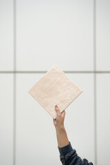 Female Hand Holding Square Beige Tile Against White Tiled Background