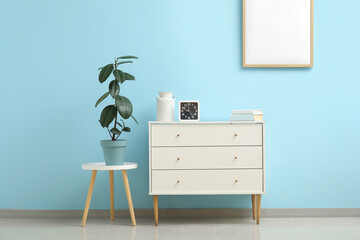 Stylish white chest of drawers with clock and houseplant on stool near blue wall