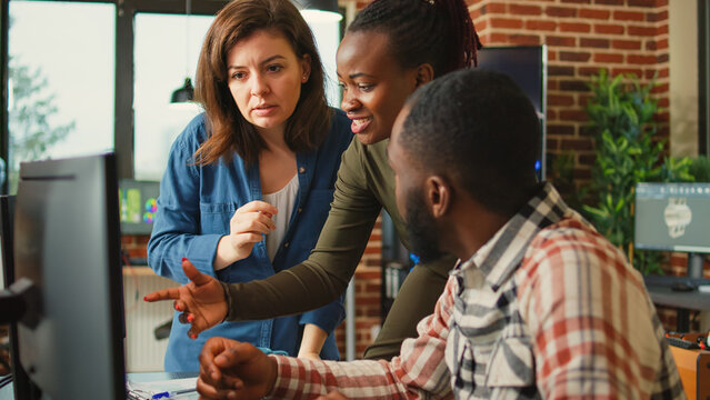 Multiethnic team of employees talking about engine component on computer, analyzing product on engineering software. Diverse colleagues checking manufacturing interface in creative studio.