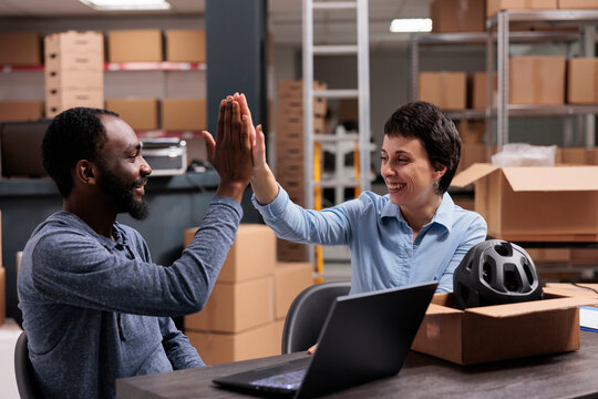 Smiling workers doing high five after putting helmet order for client in cardboard box finishing work discussing about shipping logistics. Happy team celebrating triumph in warehouse