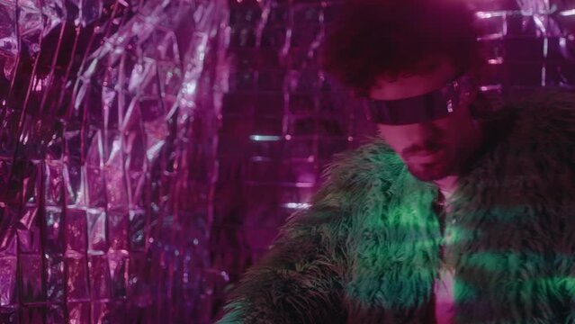 Young stylish man in futuristic monoblock sunglasses and feather jacket posing in colorful neon light against foil background. Creative studio portrait, fashion photoshoot