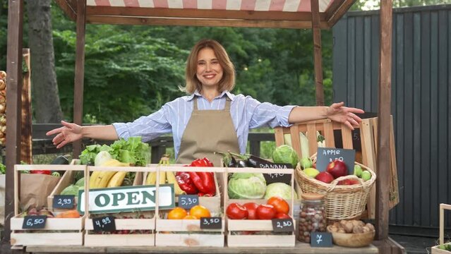 Beautiful Caucasian female farmer with apron putting on wooden desk open sign. Adorable woman standing behind stall with natural produce at local market. Positively smiling while greeting customers.
