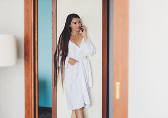 people, beauty, lifestyle, technology and relaxation concept - beautiful young woman in white bath robe with smartphone calling and talking at spa.