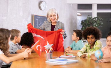 Smiling mature female teacher working in a high school tells pupils the history of Turkey in class and holds the national flag .of the country in her hands