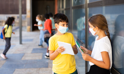 Boy and girl in face masks looking at exercise book talking about homework after classes near school