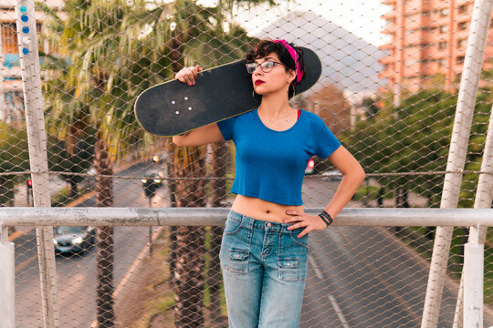bella chica patinadora con actitud y posando de pie con su skate, con maquillaje, lentes y estilo 80s 90s en la ciudad.