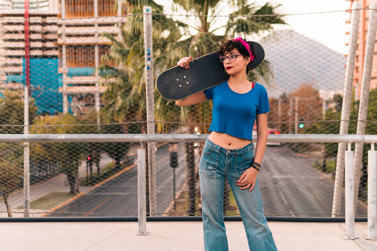 bella chica patinadora con actitud y posando de pie con su skate, con maquillaje, lentes y estilo 80s 90s en la ciudad.
